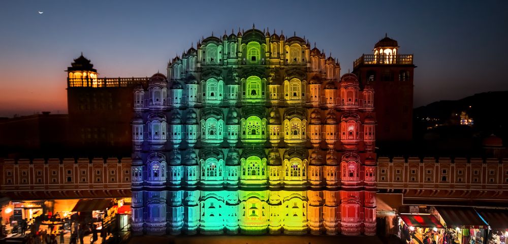Hawa Mahal illuminated in vibrant rainbow colors at dusk, with intricate windows, creating a festive, celebratory atmosphere. Silhouetted sky above.