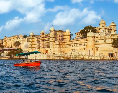 A small red boat with a green canopy floats on a lake in front of a grand, ornate palace with domed towers, under a bright blue sky with fluffy clouds.