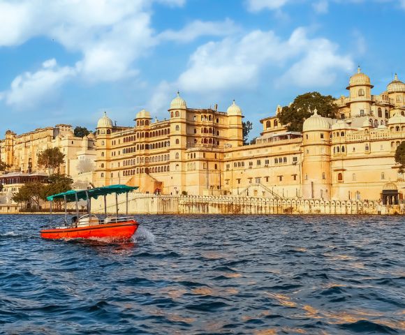 A small red boat with a green canopy floats on a lake in front of a grand, ornate palace with domed towers, under a bright blue sky with fluffy clouds.