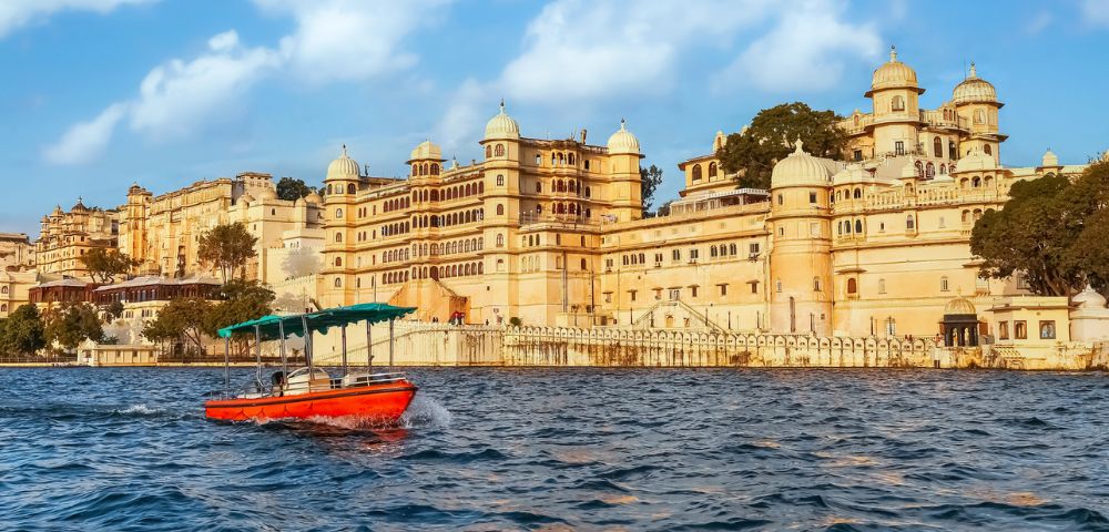 A small red boat with a green canopy floats on a lake in front of a grand, ornate palace with domed towers, under a bright blue sky with fluffy clouds.