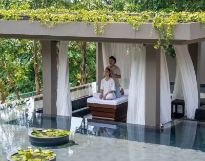 A person relaxes on a lounge chair under a canopy beside a pool surrounded by lush greenery.
