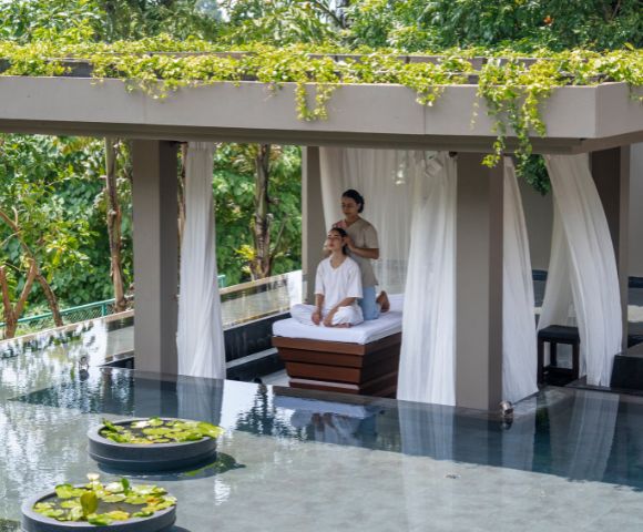 A person relaxes on a lounge chair under a canopy beside a pool surrounded by lush greenery.