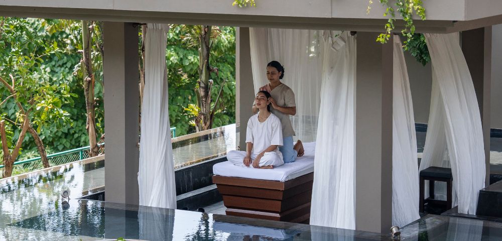 A person relaxes on a lounge chair under a canopy beside a pool surrounded by lush greenery.
