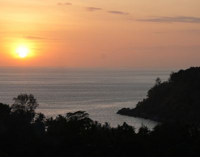 A serene sunset over calm waters, with silhouetted trees and a distant shoreline, creating a peaceful coastal scene.
