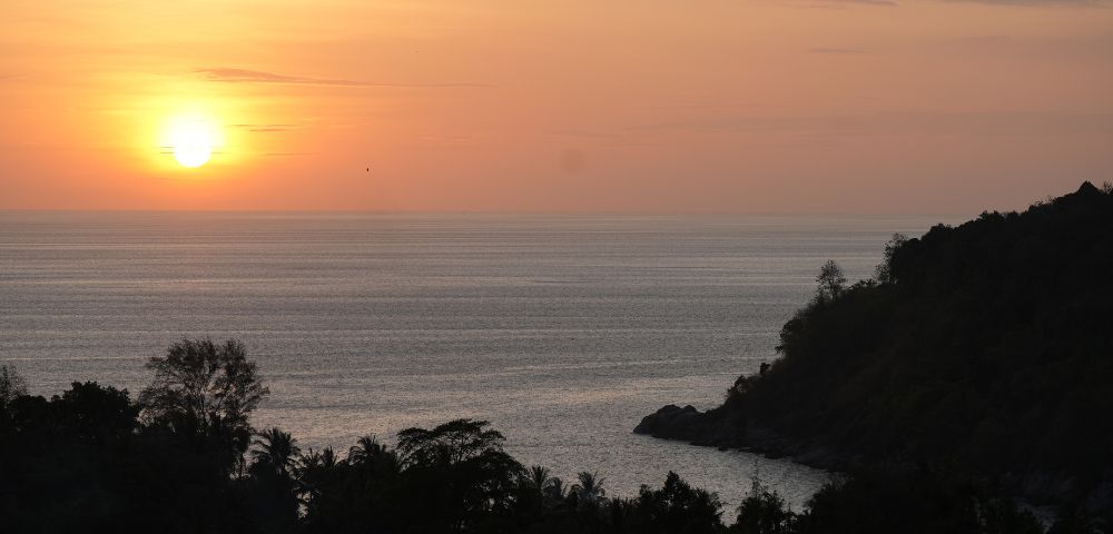 A serene sunset over calm waters, with silhouetted trees and a distant shoreline, creating a peaceful coastal scene.
