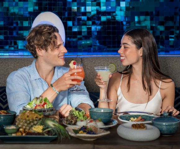Two people enjoy food and drinks at a table, smiling warmly at each other.