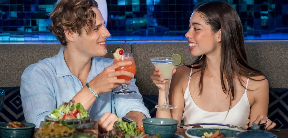 Two people enjoy food and drinks at a table, smiling warmly at each other.