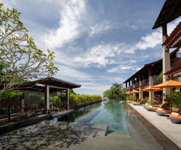 An infinity pool stretches between buildings with lounge chairs under a partly cloudy sky.
