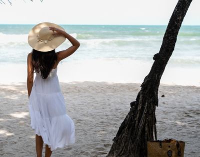 A person in a white dress and hat stands on a sandy beach, gazing at the ocean.