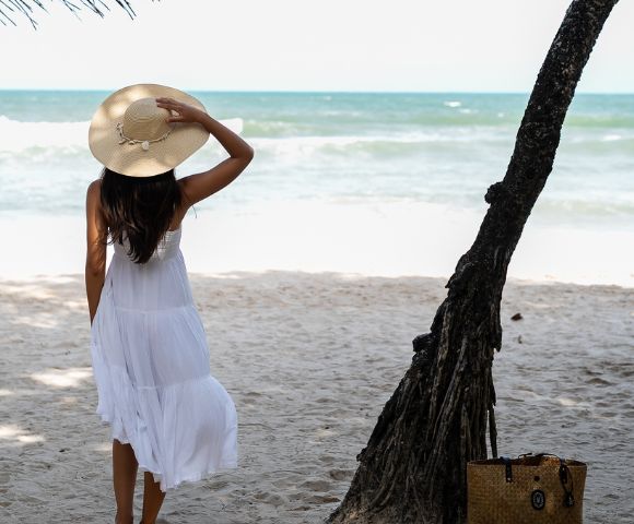 A person in a white dress and hat stands on a sandy beach, gazing at the ocean.