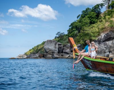 A couple relaxes on the bow of a wooden boat, surrounded by clear water and lush green cliffs under a bright blue sky.