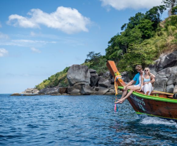 A couple relaxes on the bow of a wooden boat, surrounded by clear water and lush green cliffs under a bright blue sky.