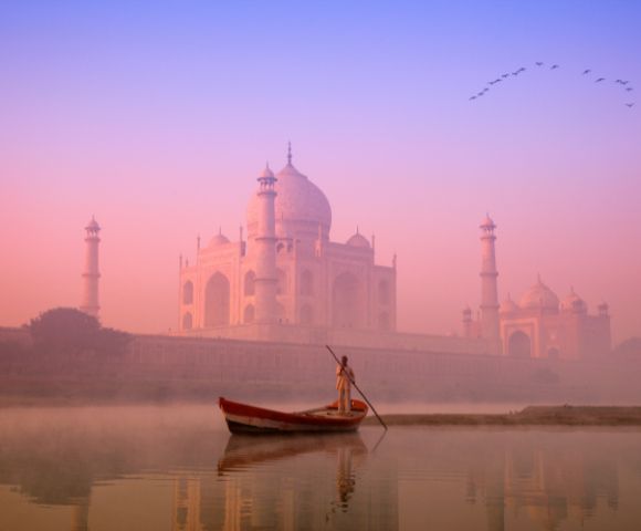 A person in a small boat rows on a calm river at sunrise, with the Taj Mahal in soft pink and orange hues and birds flying in a clear sky above.