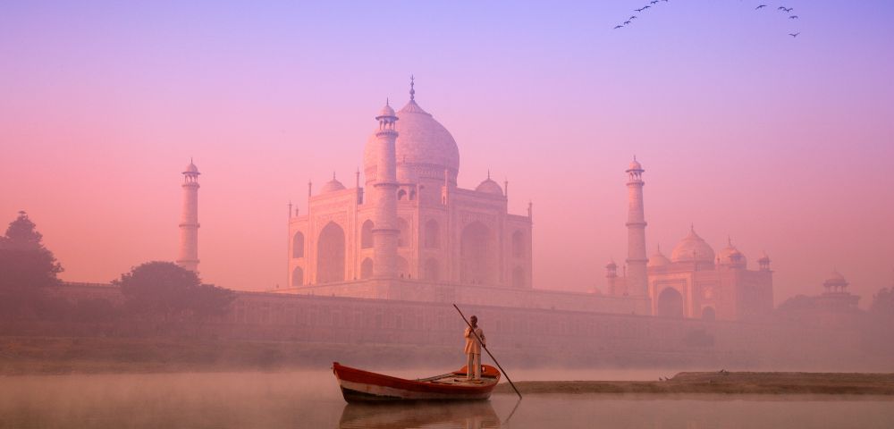 A person in a small boat rows on a calm river at sunrise, with the Taj Mahal in soft pink and orange hues and birds flying in a clear sky above.