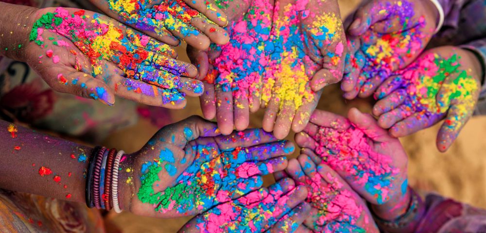 A circle of hands covered in vibrant colored powders, including pink, yellow, blue, and green, conveying a festive and joyful atmosphere.