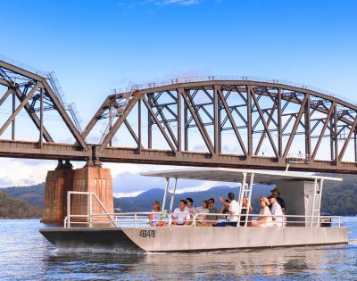 A steel truss bridge spans a calm river with a boat passing underneath, set against a clear blue sky.