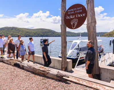 A group of people gather near the shore of a lake or sea, with mountains in the background, boats docked nearby, and a wooden signpost with an unreadable sign.