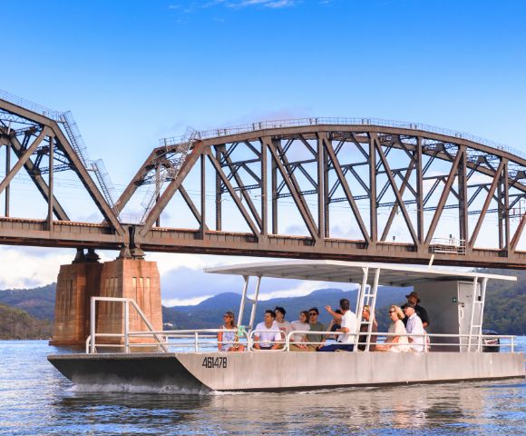 A steel truss bridge spans a calm river with a boat passing underneath, set against a clear blue sky.