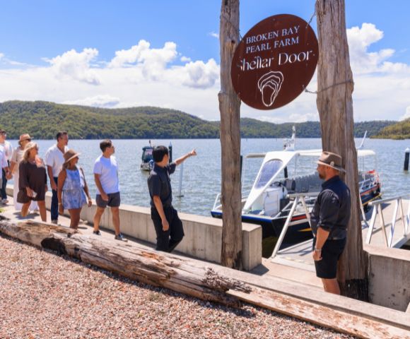 A group of people gather near the shore of a lake or sea, with mountains in the background, boats docked nearby, and a wooden signpost with an unreadable sign.