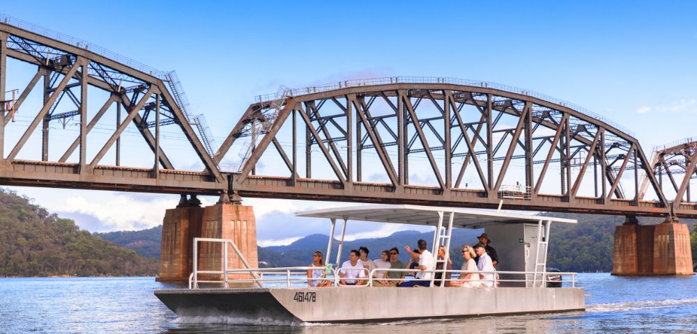 A steel truss bridge spans a calm river with a boat passing underneath, set against a clear blue sky.