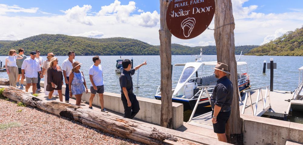 A group of people gather near the shore of a lake or sea, with mountains in the background, boats docked nearby, and a wooden signpost with an unreadable sign.