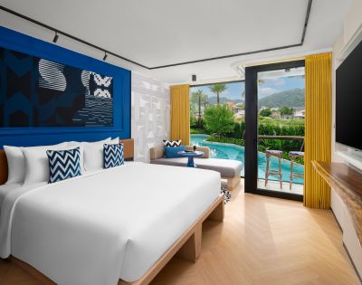 A modern hotel room with a large bed, blue accent wall, and sliding glass doors opening to a balcony with outdoor seating.