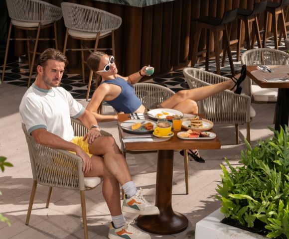 Two people seated at an outdoor café table, enjoying drinks and food under a shaded area surrounded by greenery.