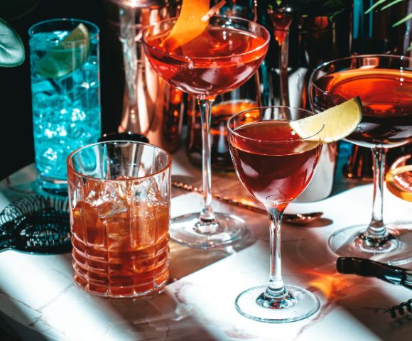 Close-up of several colorful cocktails on a bar counter, including red and orange drinks garnished with fruit slices.