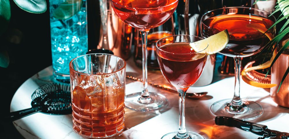 Close-up of several colorful cocktails on a bar counter, including red and orange drinks garnished with fruit slices.