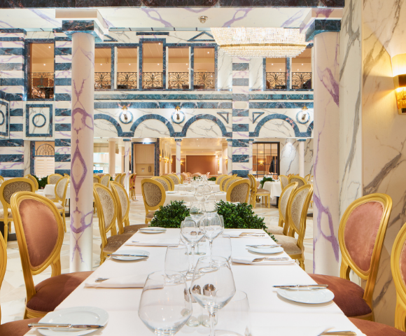 Elegant restaurant interior featuring a long table set with glassware, surrounded by ornate gold chairs and marble columns.