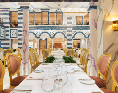 Elegant restaurant interior featuring a long table set with glassware, surrounded by ornate gold chairs and marble columns.