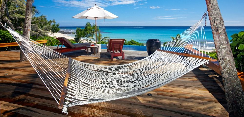 A serene beachside scene featuring a hammock on a wooden deck, overlooking turquoise waters and a sunny sky with a parasol.