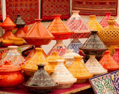 Colorful Moroccan tagine pots with intricate patterns are displayed at a market. They are vibrant in orange, yellow, red, and black, creating a lively, cultural scene.