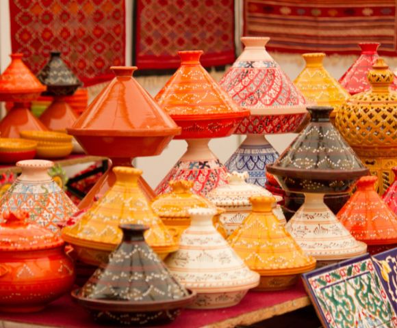 Colorful Moroccan tagine pots with intricate patterns are displayed at a market. They are vibrant in orange, yellow, red, and black, creating a lively, cultural scene.