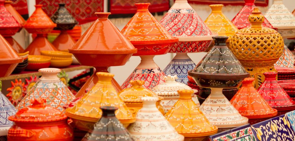Colorful Moroccan tagine pots with intricate patterns are displayed at a market. They are vibrant in orange, yellow, red, and black, creating a lively, cultural scene.