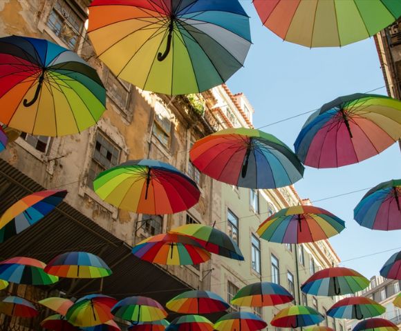 Colorful umbrellas suspended above a narrow street, creating a vibrant canopy against a clear blue sky. The scene conveys a playful and cheerful atmosphere.