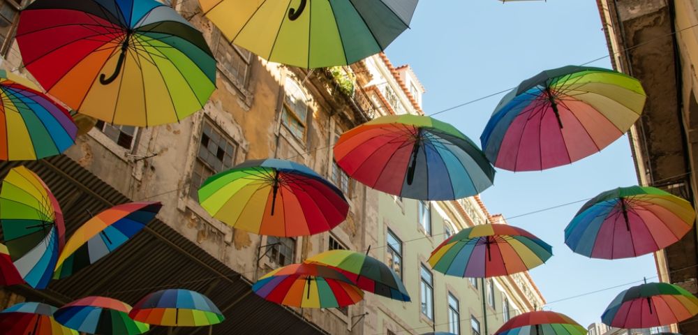Colorful umbrellas suspended above a narrow street, creating a vibrant canopy against a clear blue sky. The scene conveys a playful and cheerful atmosphere.