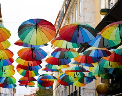 Colorful umbrellas hang above an urban street, creating a vibrant canopy. The scene is lively and artistic, with bright hues against a building backdrop.