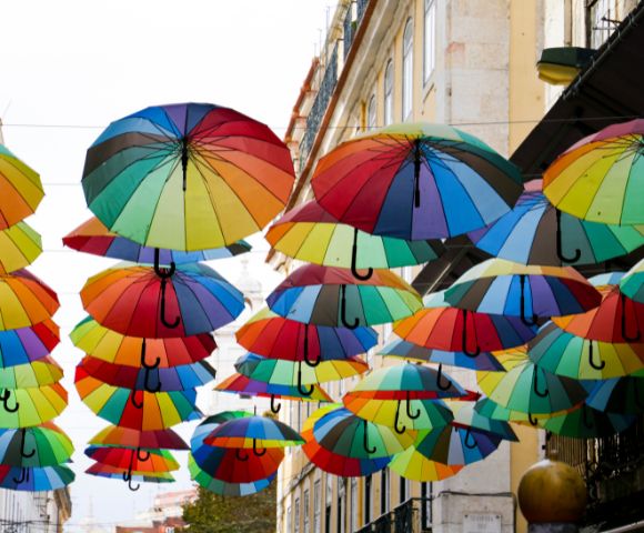 Colorful umbrellas hang above an urban street, creating a vibrant canopy. The scene is lively and artistic, with bright hues against a building backdrop.