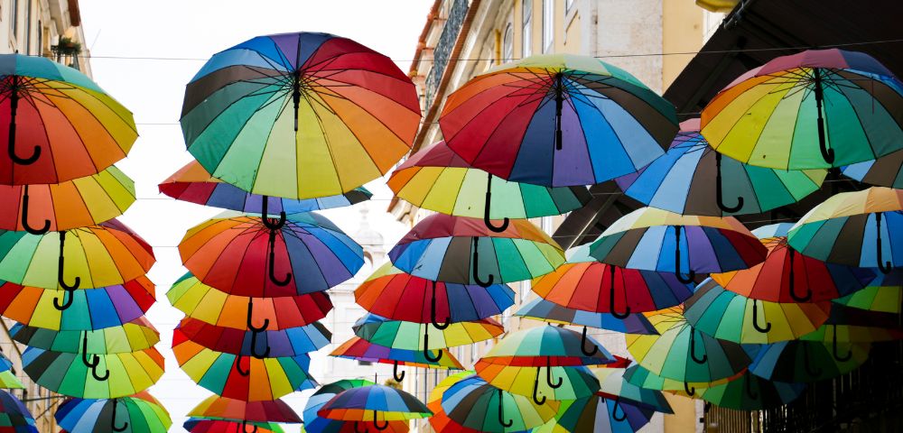 Colorful umbrellas hang above an urban street, creating a vibrant canopy. The scene is lively and artistic, with bright hues against a building backdrop.