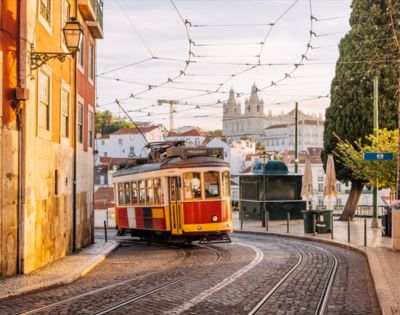 A classic yellow and red tram curves along cobblestone streets in a European city. Historic buildings and a cathedral tower are visible under a sunny sky.