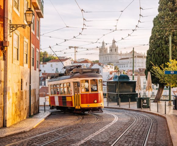 A classic yellow and red tram curves along cobblestone streets in a European city. Historic buildings and a cathedral tower are visible under a sunny sky.