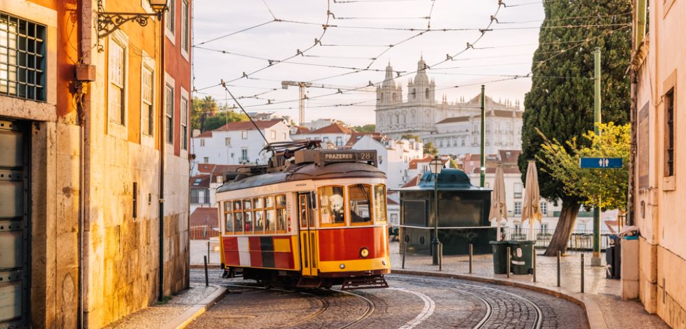 A classic yellow and red tram curves along cobblestone streets in a European city. Historic buildings and a cathedral tower are visible under a sunny sky.