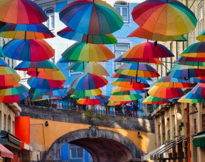 Colorful umbrellas hang above a lively street, creating a vibrant canopy. The mood is cheerful and whimsical, with bright buildings in the background.