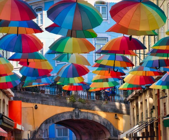 Colorful umbrellas hang above a lively street, creating a vibrant canopy. The mood is cheerful and whimsical, with bright buildings in the background.