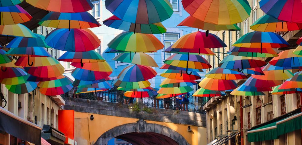 Colorful umbrellas hang above a lively street, creating a vibrant canopy. The mood is cheerful and whimsical, with bright buildings in the background.