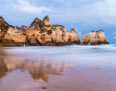 Rocky cliffs reflected in calm ocean water under a cloudy sky at dusk. The serene landscape exudes tranquility, with gentle waves lapping at the shore.