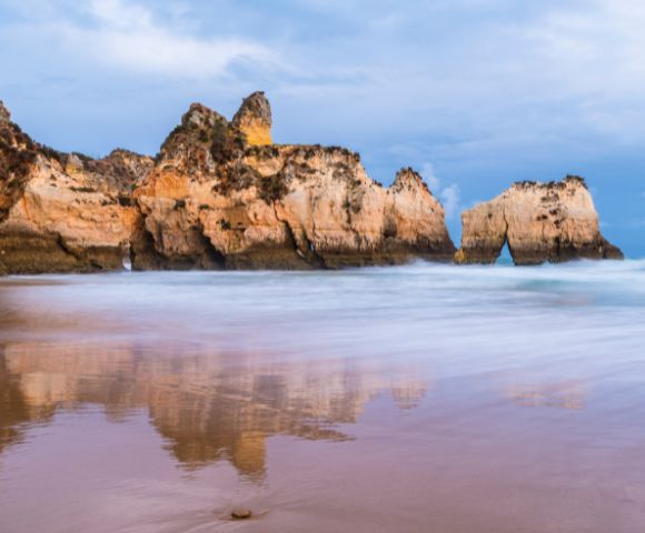 Rocky cliffs reflected in calm ocean water under a cloudy sky at dusk. The serene landscape exudes tranquility, with gentle waves lapping at the shore.