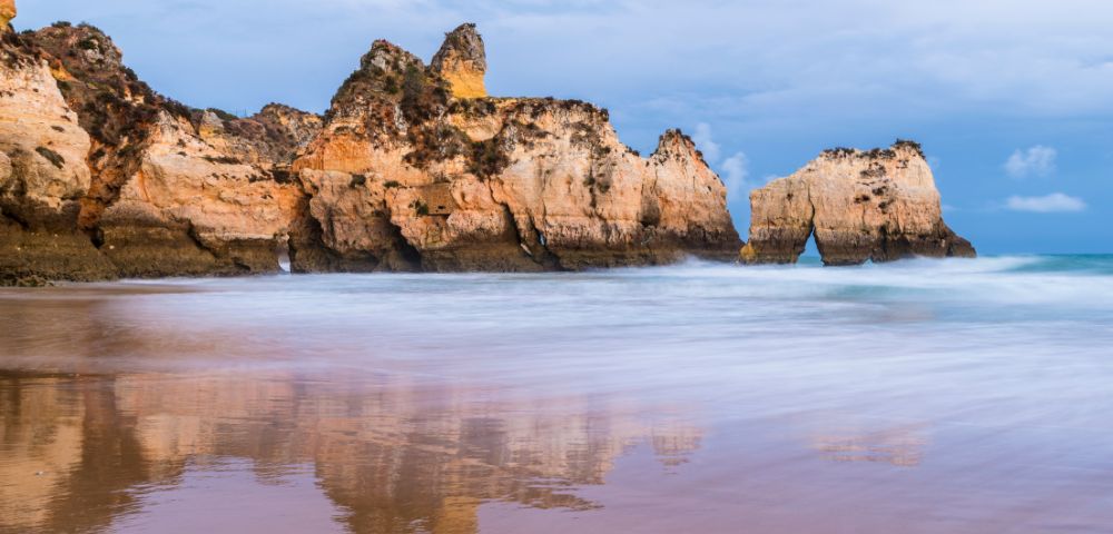 Rocky cliffs reflected in calm ocean water under a cloudy sky at dusk. The serene landscape exudes tranquility, with gentle waves lapping at the shore.