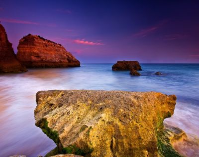 A large, textured rock dominates the foreground against a blurred seascape at dusk, with vibrant purple and orange skies and distant cliffs enhancing tranquility.